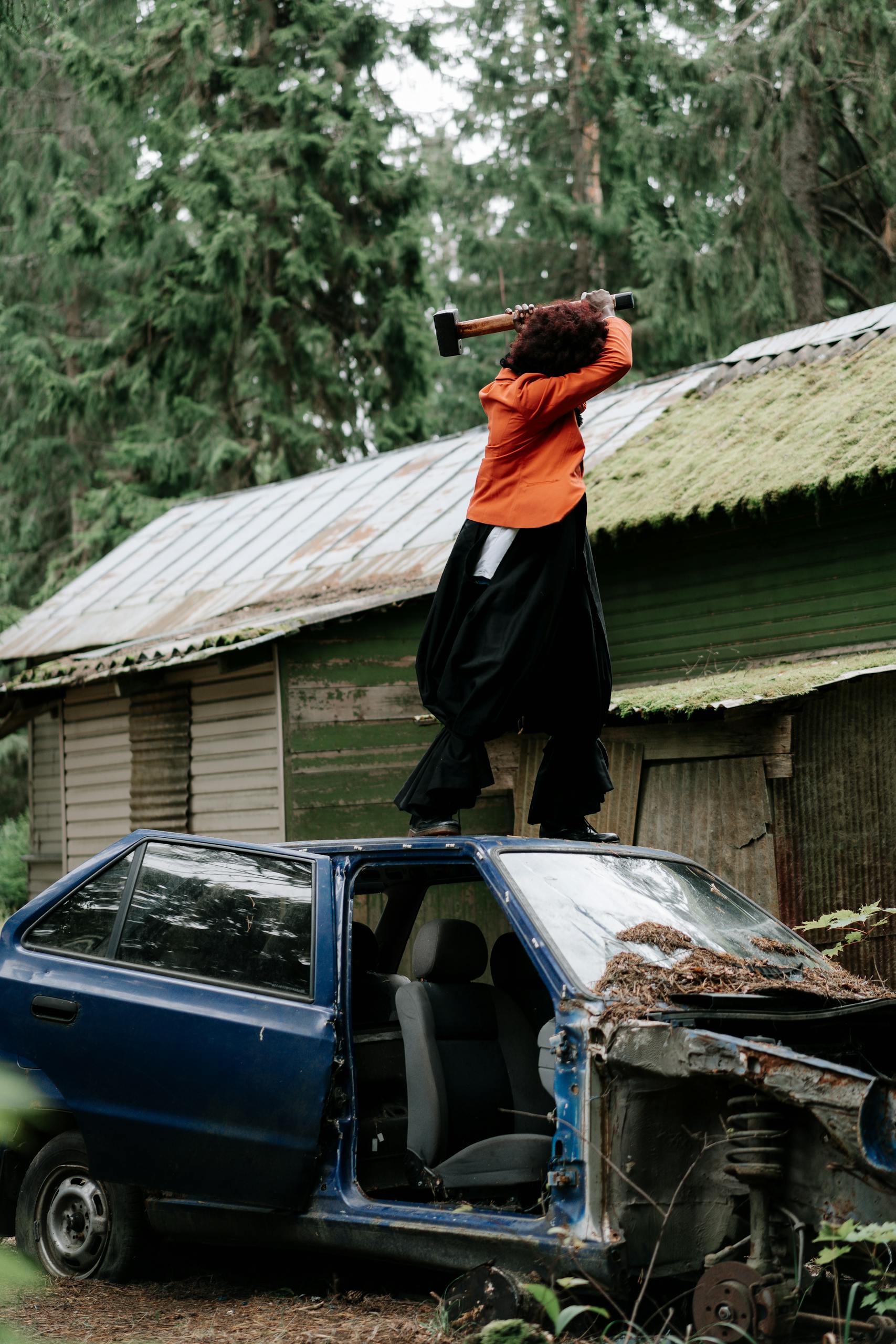 A dramatic scene of a person with a hammer smashing an abandoned car in a spooky forest setting.